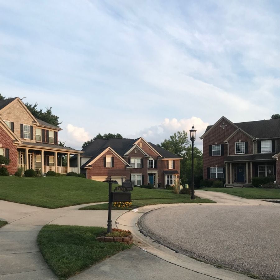 A quiet suburban street lined with large, two-story brick houses, neatly manicured lawns, and a curved sidewalk. A black mailbox with flowers is visible in the foreground under a partly cloudy sky.