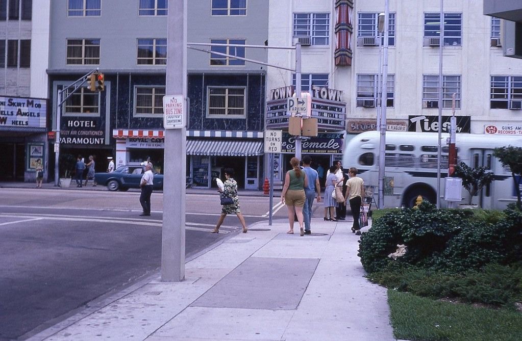 People walk along a city sidewalk near a street corner with a bus passing by. There are shops, a hotel, and a theater with a vintage marquee in the background. Some traffic signs and greenery are visible.