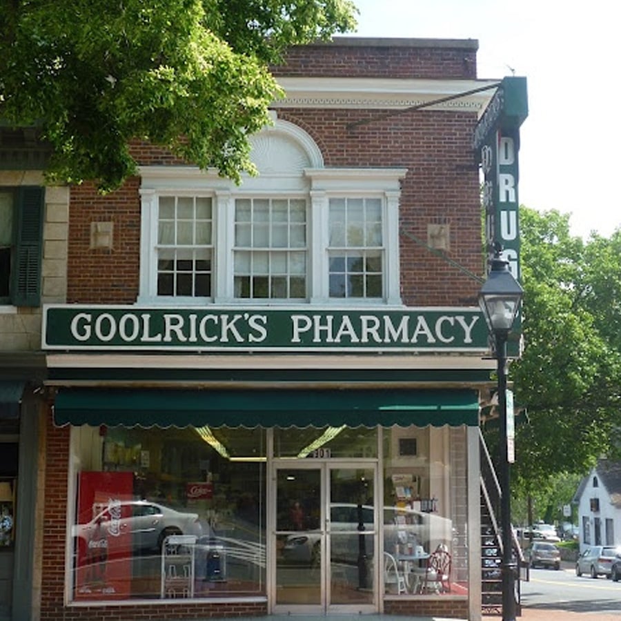 A brick building with a green awning and a large sign reading "Goolrick's Pharmacy." The entrance has glass doors, and a vertical sign on the side reads "DRUGS." Trees and cars are visible nearby.
