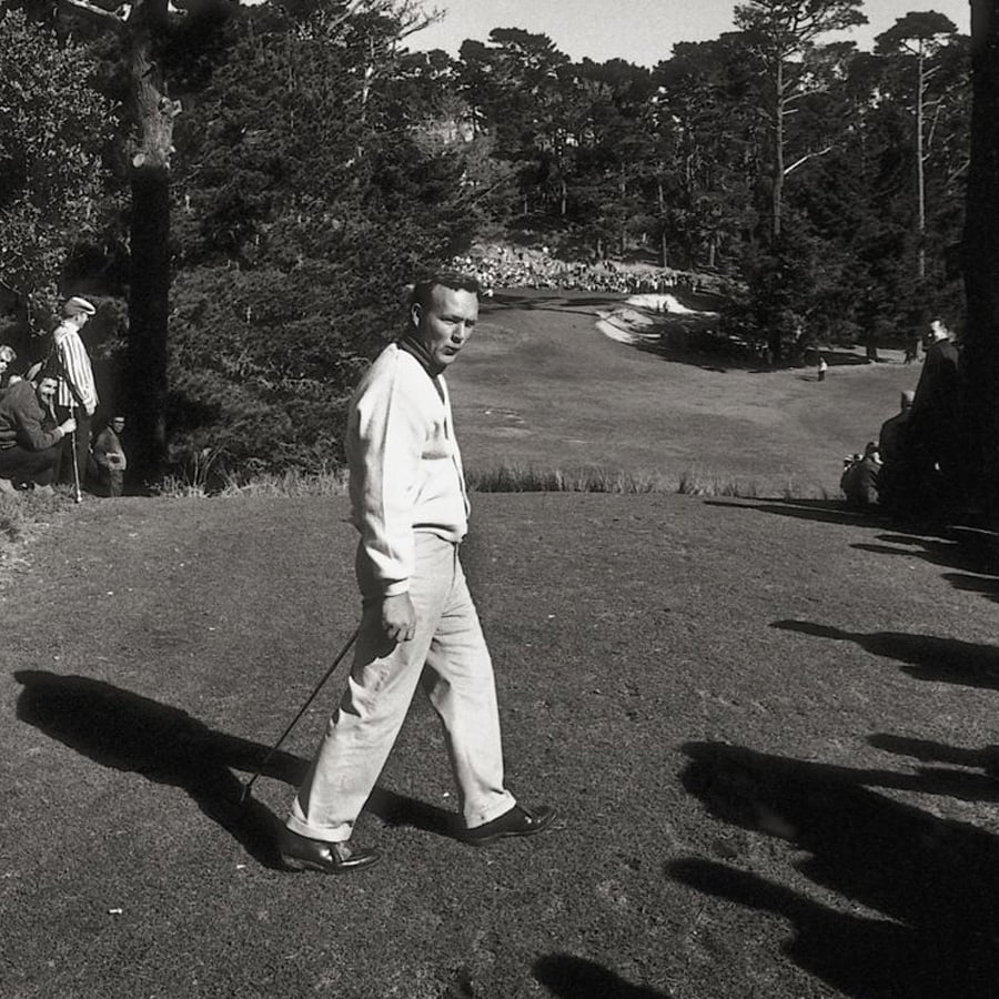 A man in golf attire stands on a golf course holding a club, looking back over his shoulder. Spectators line both sides, and tall trees surround the area under a clear sky. The scene appears to be from decades past.