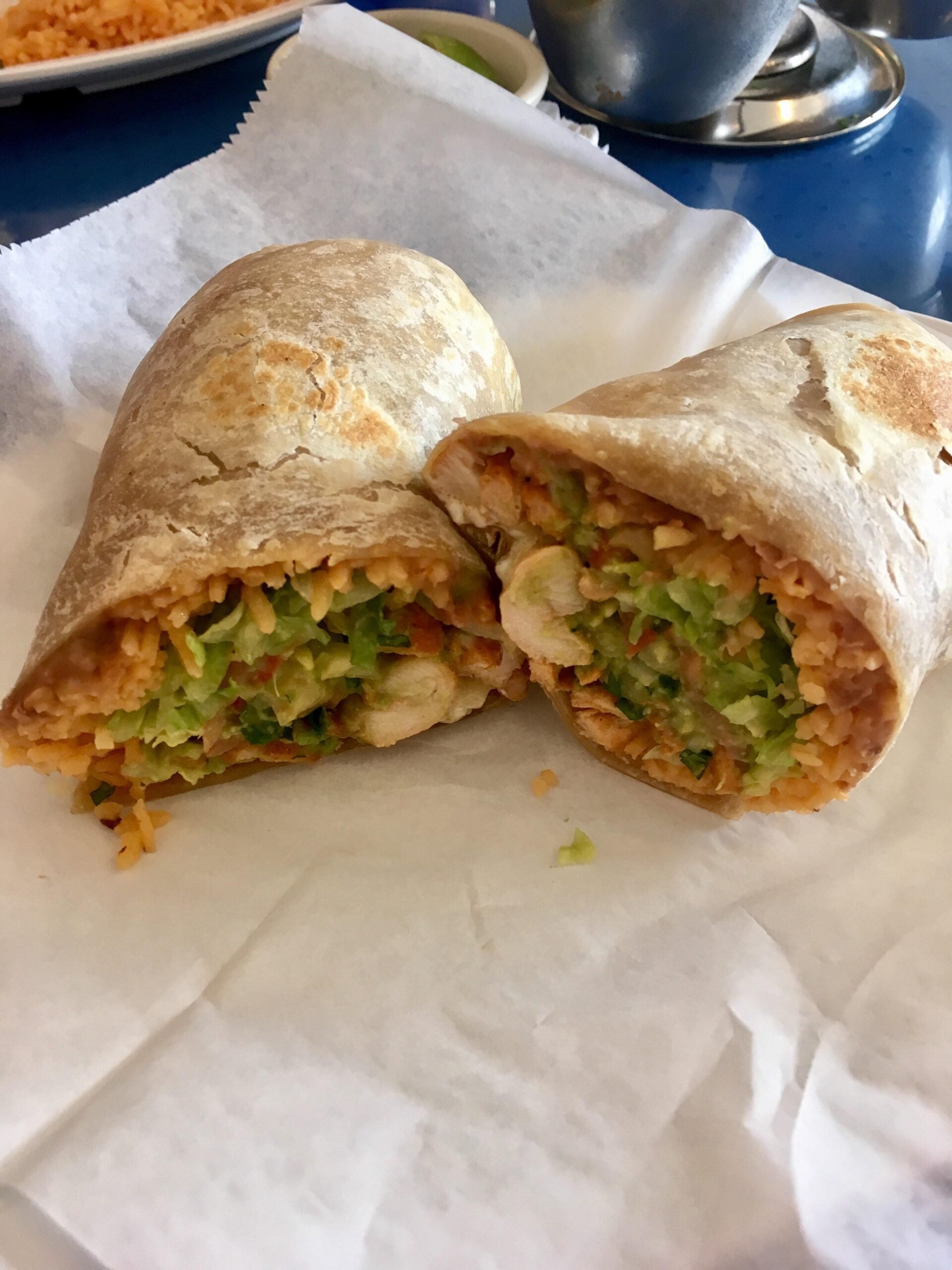 A burrito cut in half, showing a filling of rice, lettuce, beans, and chicken, placed on white parchment paper. In the background, a plate of rice and a metal sauce container are partially visible.
