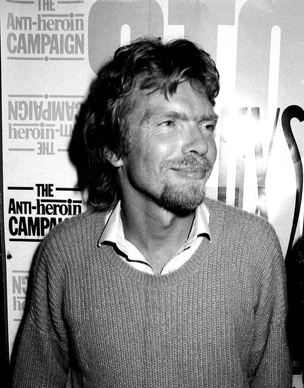 A man with medium-length hair and a beard, wearing a light sweater over a collared shirt, stands in front of a poster with the words "The Anti-heroin Campaign" visible on it. The photo is in black and white.