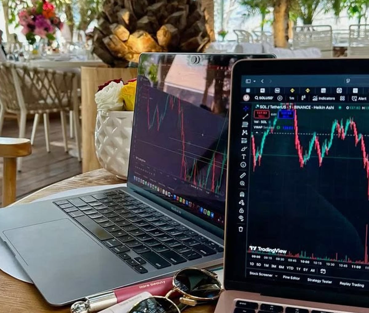 Two open laptops display cryptocurrency trading charts at an outdoor café. The table also holds sunglasses, a pen, and a vase of flowers. Palm trees and seating are visible in the background.