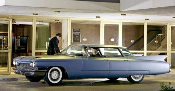 A vintage blue Cadillac is parked under the entrance of a modernist building at night. A man stands beside the car, while another person sits inside. The building features large glass windows and geometric architecture.