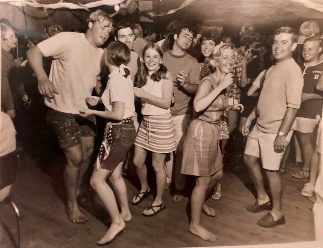 A group of young adults dance and smile at a lively indoor party, dressed in casual summer clothes. Streamers hang from the ceiling and people are mingling in the background. The photo is in black and white.