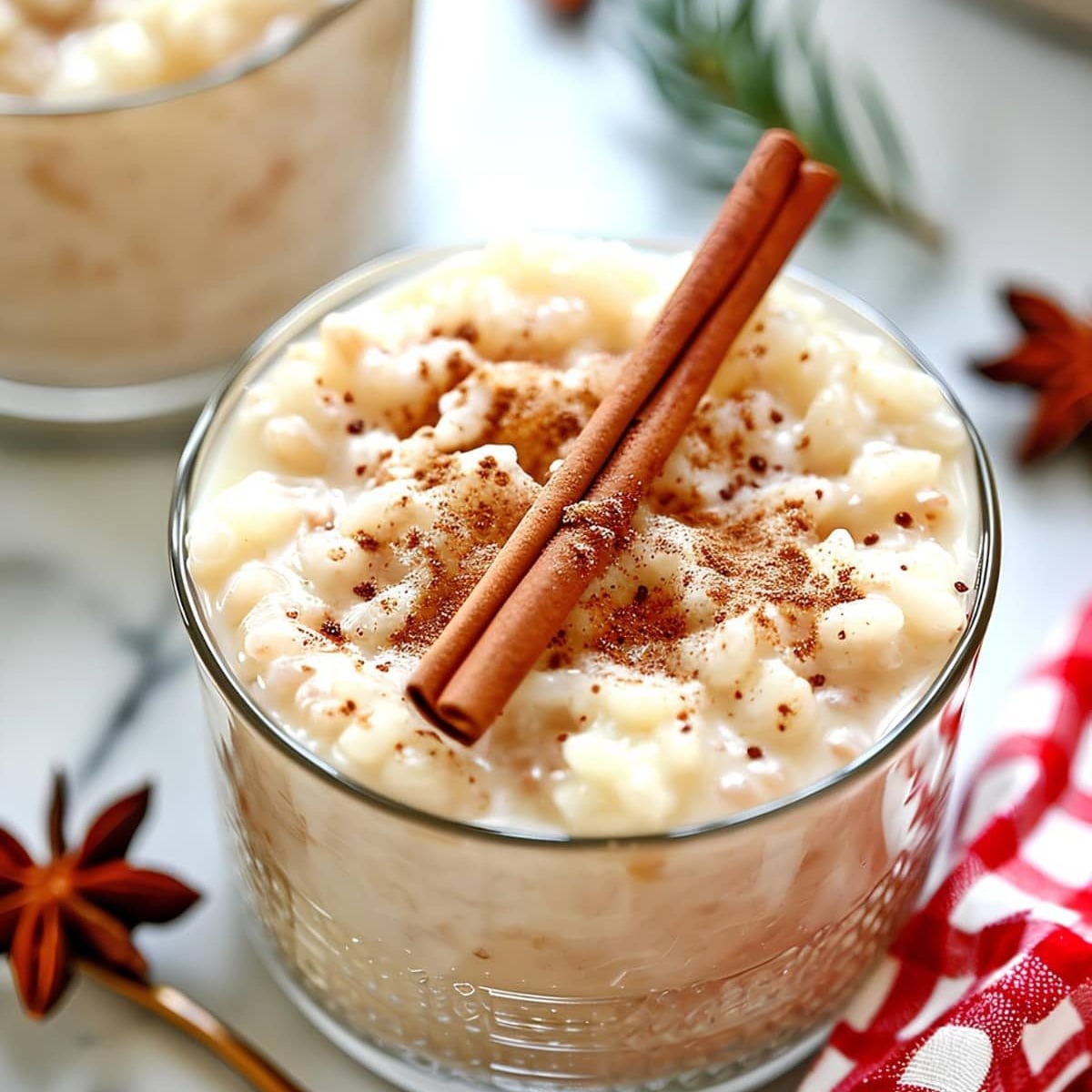 A glass filled with creamy rice pudding, topped with ground cinnamon and two cinnamon sticks. Anise stars and a red checkered cloth are nearby, creating a cozy, inviting presentation.