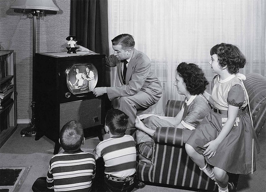 A black-and-white photo shows a family gathered in a living room, watching an old-fashioned television. A man points at the TV screen while two children sit on the floor and a woman and girl sit on a couch, all focused on the screen.