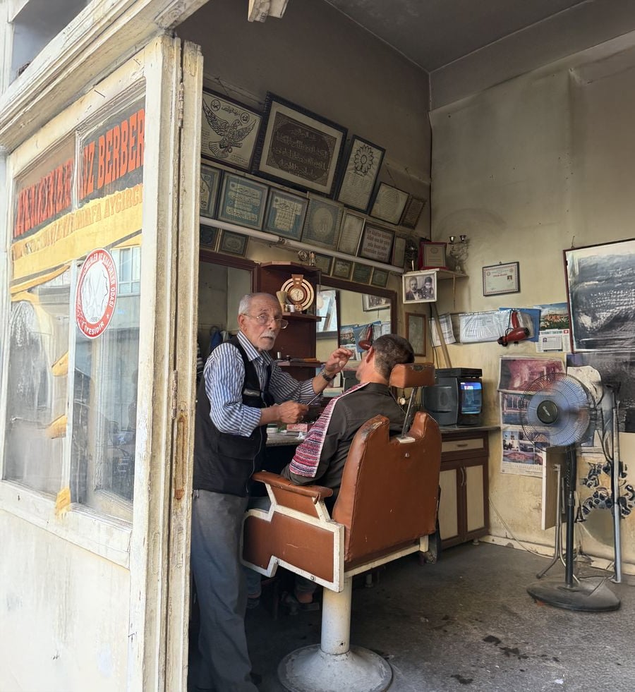 An elderly barber trims a customer’s beard inside a small, vintage barbershop filled with framed certificates and old photographs on the walls. Sunlight streams in through a window with a yellowed shop sign.