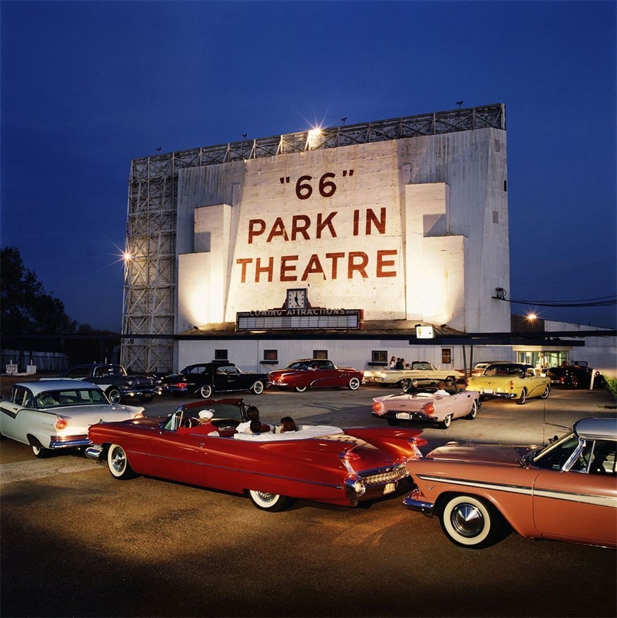 Classic cars parked at a retro drive-in theater at dusk, with people sitting in their vehicles facing a large screen reading “66 Park In Theatre.” The scene is brightly lit, evoking a nostalgic, vintage atmosphere.