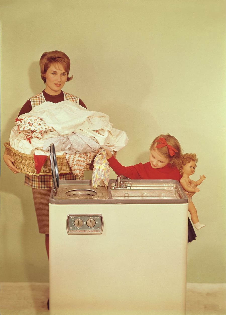 A woman holding a large laundry basket stands beside a washing machine, while a young girl holding a doll helps load clothes into the machine. The background is plain and light-colored.
