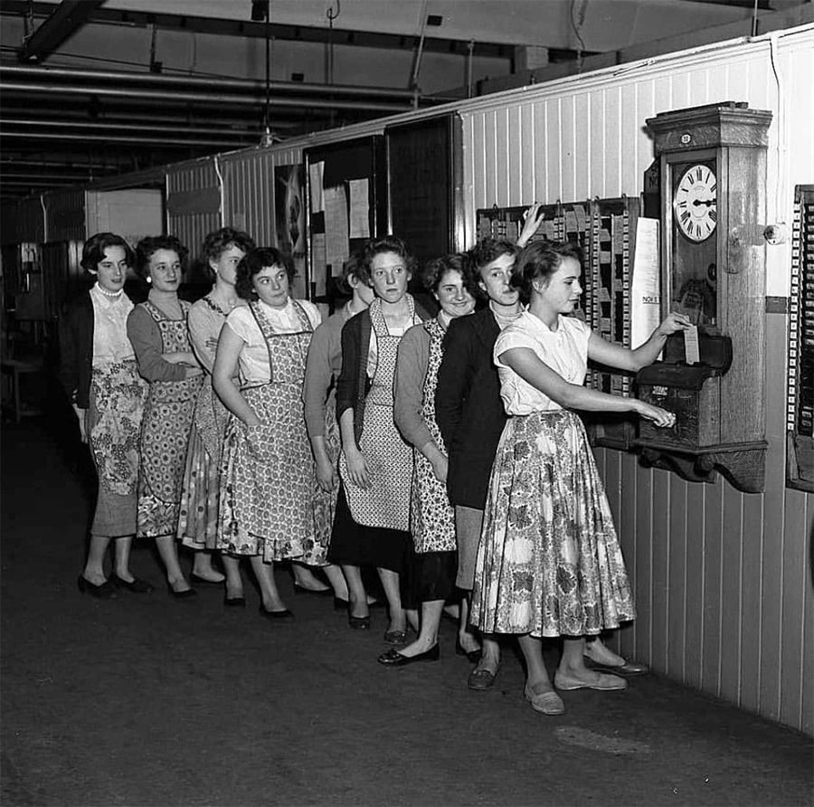 A black and white photo of women in dresses and aprons standing in a line, waiting to use a wall-mounted clocking-in machine at work, likely in a factory setting.