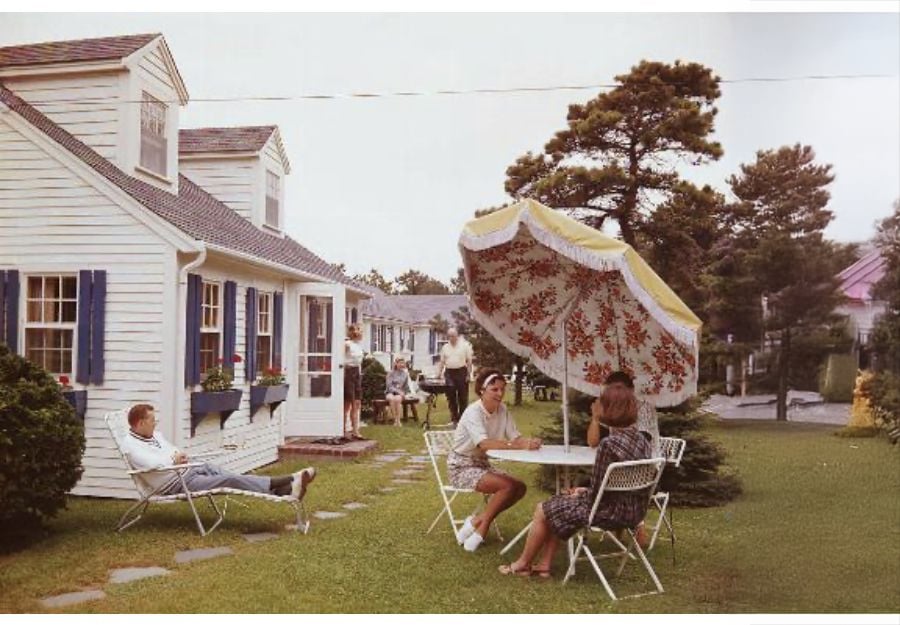 Four people relax in lawn chairs around a table under a large floral-patterned umbrella outside a white house with blue shutters; another person stands in the background amid greenery.