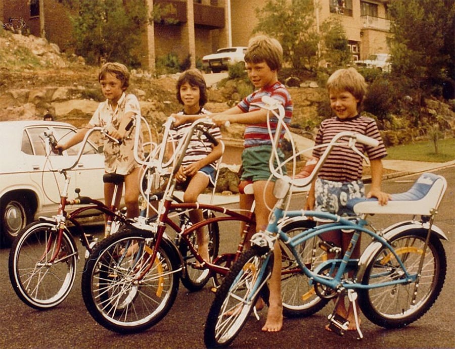 Four children stand outdoors with vintage bicycles, smiling at the camera. They are on a residential street with houses and a white car in the background. The scene appears to be from the 1970s or 1980s.