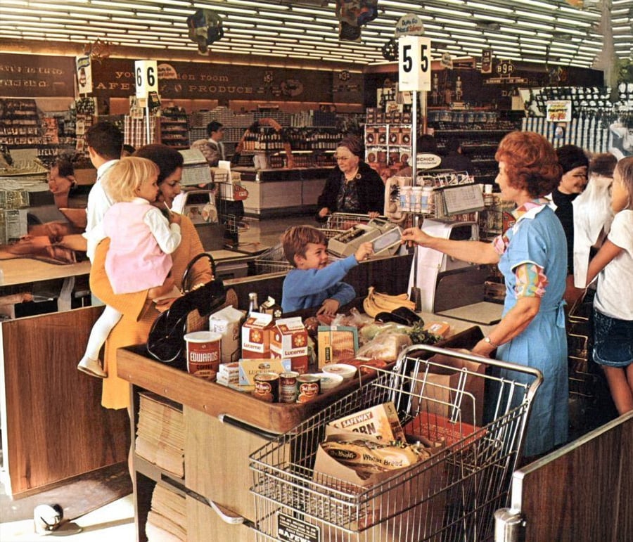A busy 1970s supermarket checkout scene with women and children waiting in line, groceries on the counters, and a cashier handing change to a young boy. Shopping carts are filled with food items.