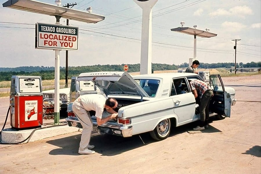 Two men examine the trunk and interior of a light blue vintage car at a Texaco gas station, with gas pumps and a sign that reads "Texaco Gasolines Localized For You" visible in the background.