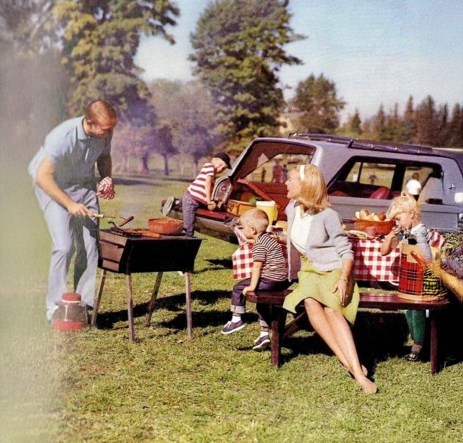 A family enjoys a picnic beside a car in a grassy park. A man grills food, while a woman and three children sit at a table with a checkered tablecloth, surrounded by trees and picnic supplies.