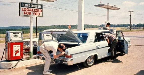 Three people are checking a white vintage car at a rural gas station with old-style pumps. The car&rsquo;s trunk and doors are open, and a sign reads &ldquo;TEXACO GASOLINES LOCALIZED FOR YOU.&rdquo;