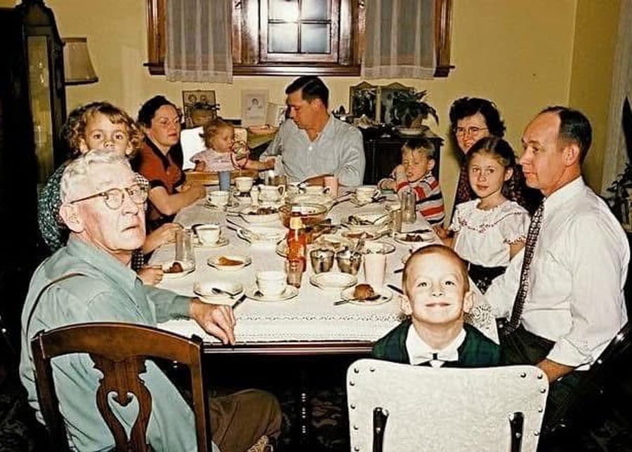 A family of ten, including young children and adults, sits around a dining table set with dishes and food, sharing a meal in a warmly lit room. All eyes are on the camera, and some are smiling.