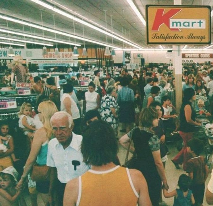 A crowded Kmart store with shoppers walking through aisles. The vintage photo shows adults and children, a retro sign reading “Kmart Satisfaction Always,” and busy checkout counters in a brightly lit space.