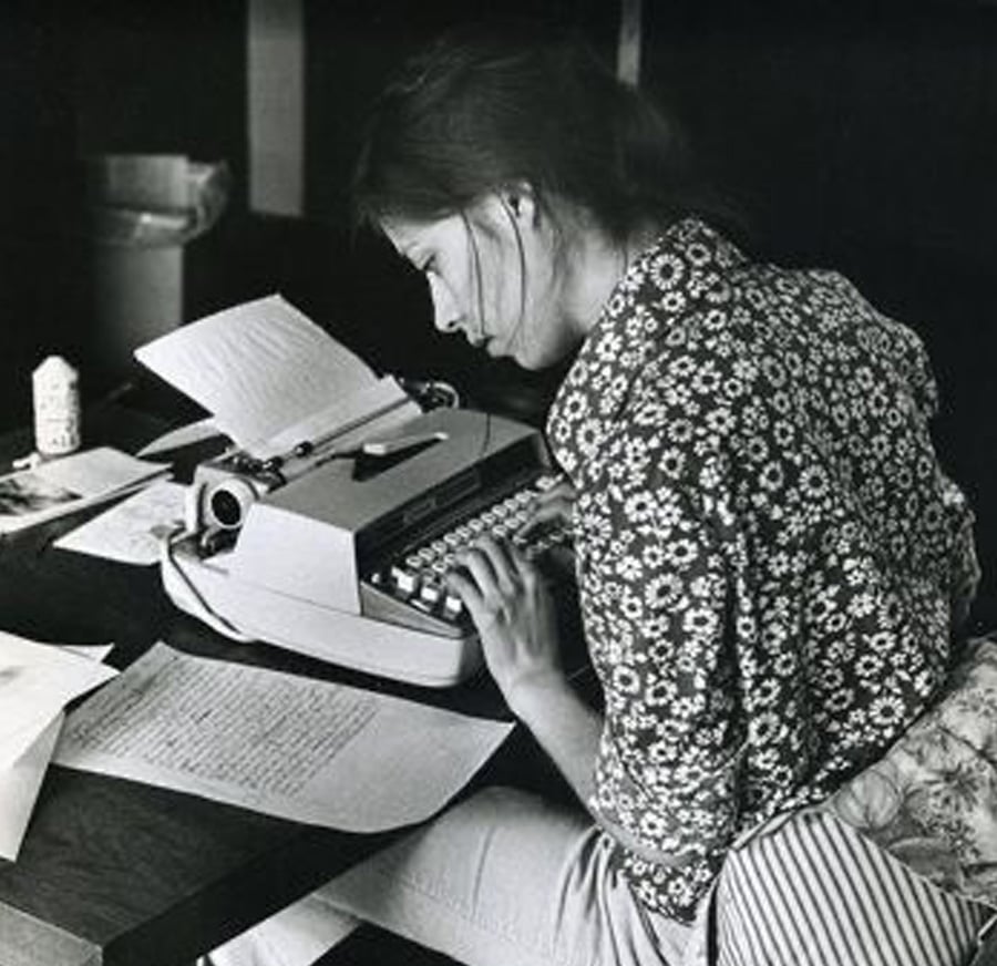 A woman sits at a desk typing on a typewriter, surrounded by scattered papers. She wears a floral-patterned shirt and appears focused on her work in a black-and-white setting.