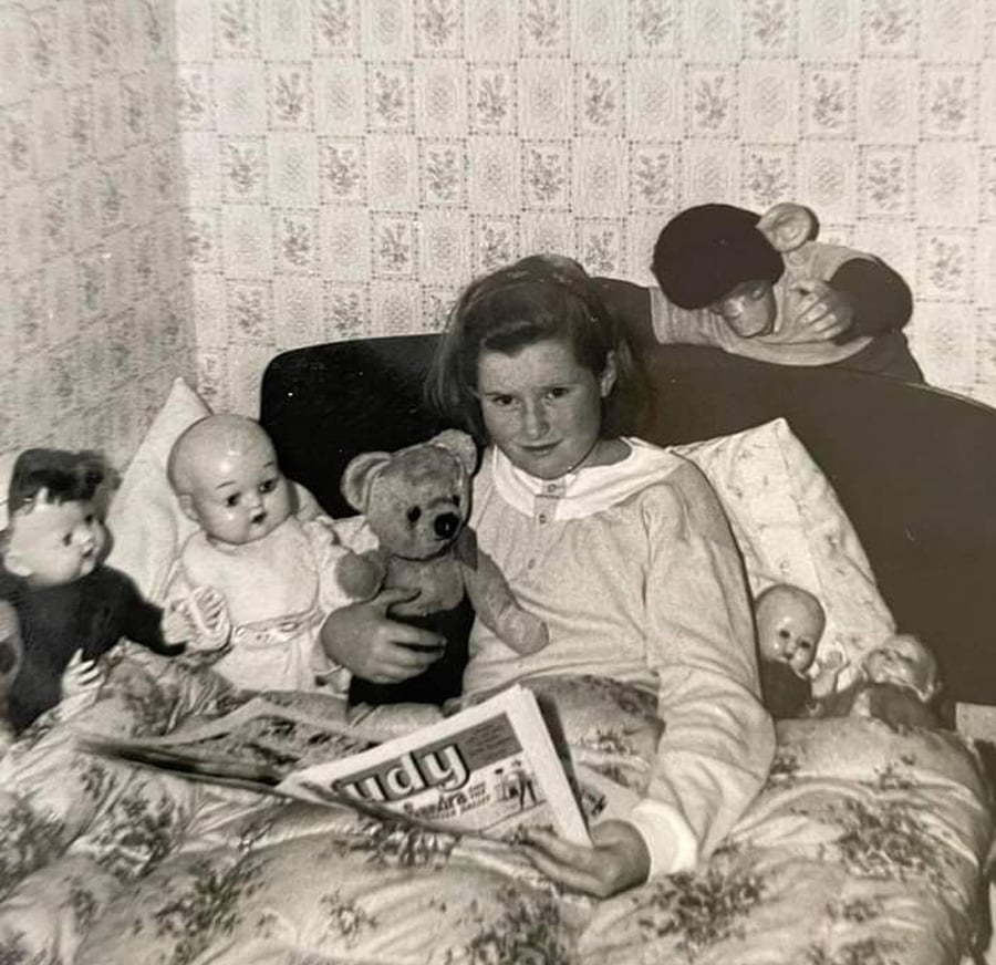A young girl sits in bed holding a teddy bear and a magazine, surrounded by dolls and stuffed toys. The room has patterned wallpaper and the bedding features a floral print. The photo is in black and white.