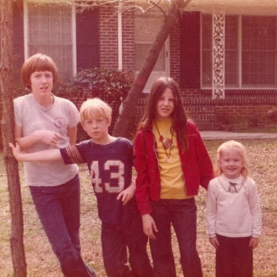 Four children stand together outside in front of a brick house. One leans on a tree, two stand close together, and the smallest child stands on the right, all posing for the photo on a grassy lawn.