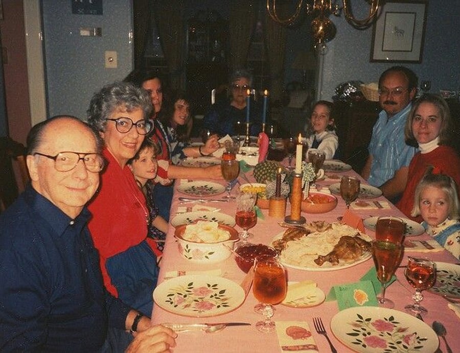 A family of eleven, spanning three generations, sits around a dining table set with food, drinks, and candles. Everyone is smiling at the camera in a warmly lit room decorated for a festive meal.