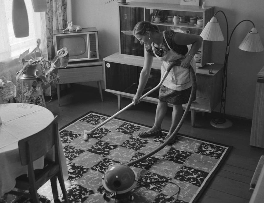 A woman in an apron vacuums a patterned rug in a vintage living room with a round table, old television, houseplants, and mid-century furniture.
