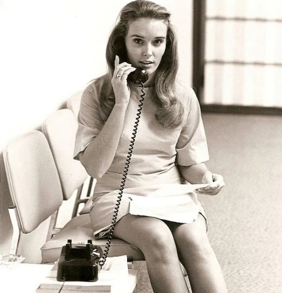 A young woman with long hair sits on a row of chairs, holding a corded telephone to her ear and a piece of paper in her other hand. She wears a short dress and looks toward the camera. A rotary phone sits on a small table nearby.