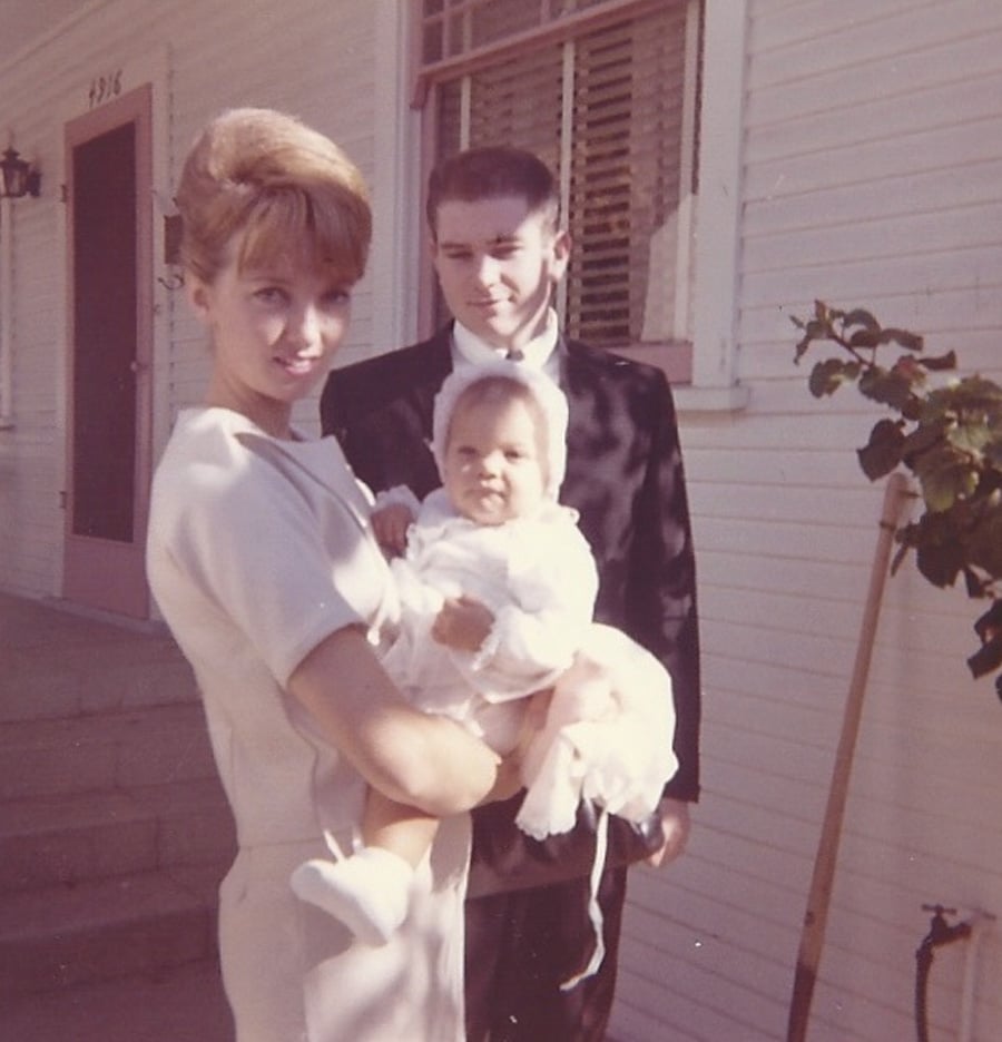 A woman in a light dress holds a baby dressed in white while a man in a dark suit stands behind them; they are outside in front of a white house with steps and a pink door.