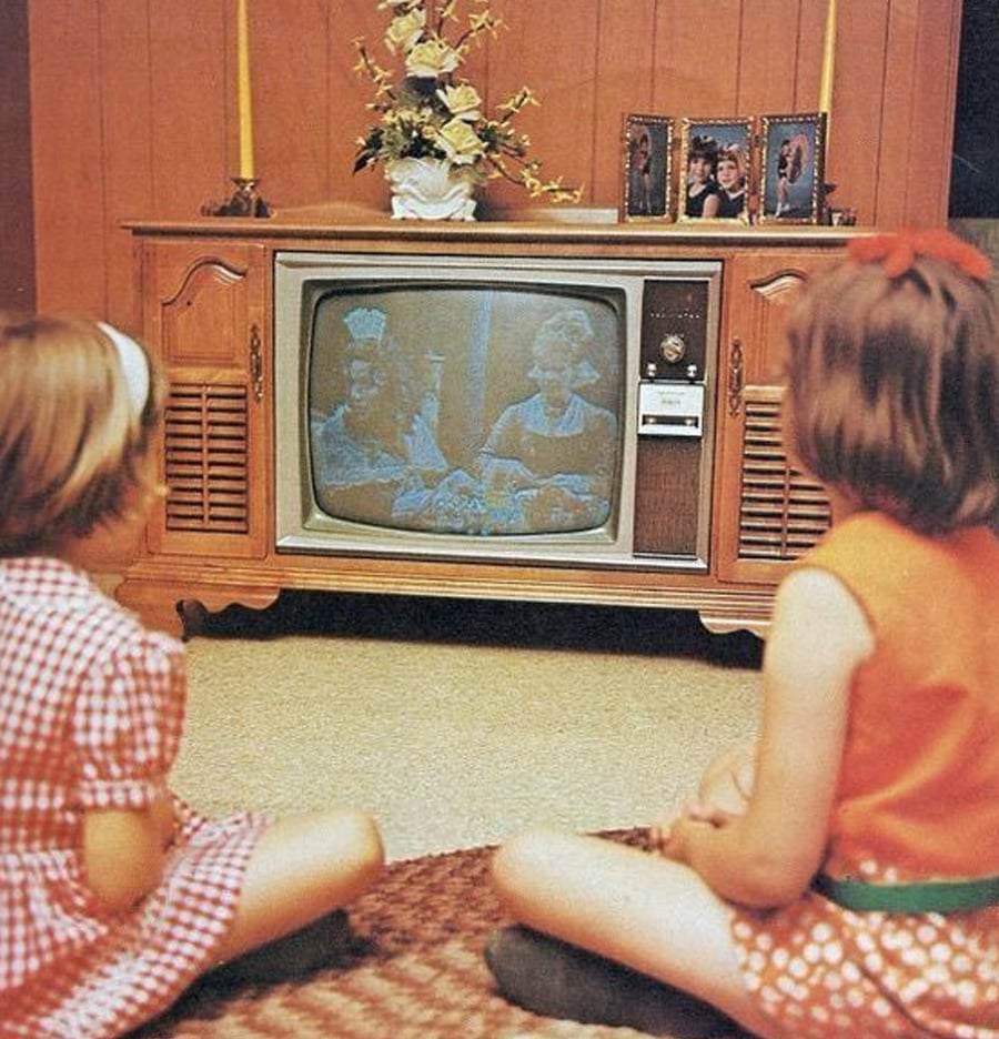 Two young girls sit on the floor watching a vintage television. The TV shows a black-and-white program. The TV is in a wooden cabinet, with candles, flowers, and framed photos on top. The room has wood-paneled walls.