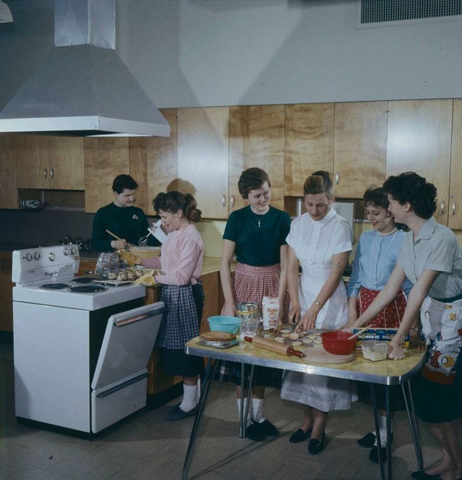 Six women in vintage clothing cook and prepare food together in a 1950s-style kitchen, smiling and working at two tables with bowls, utensils, and ingredients. A stove and wooden cabinets are in the background.