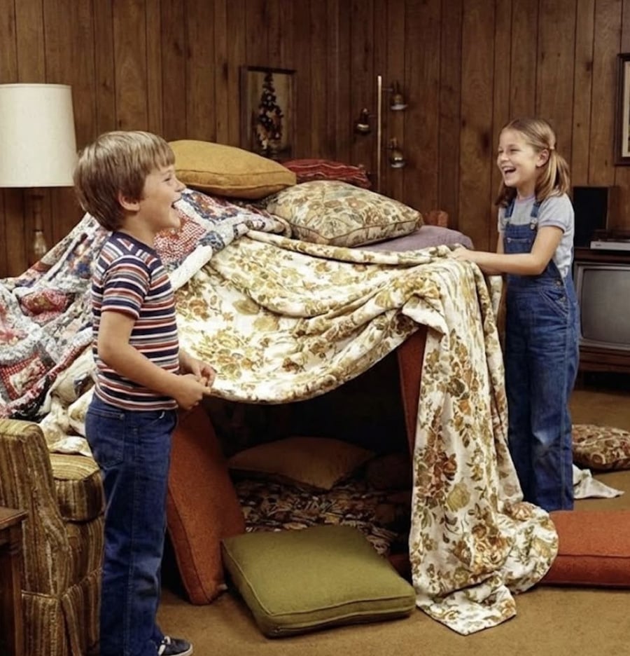 Two children smile while building a large blanket fort in a cozy, wood-paneled living room, surrounded by cushions and pillows. The scene feels playful and nostalgic.