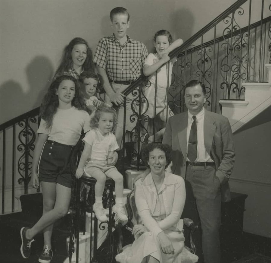 A black-and-white photo of a family with seven members posing on a staircase. Four girls, two boys, and a man in a suit stand or sit around a woman seated at the bottom, all smiling for the camera.