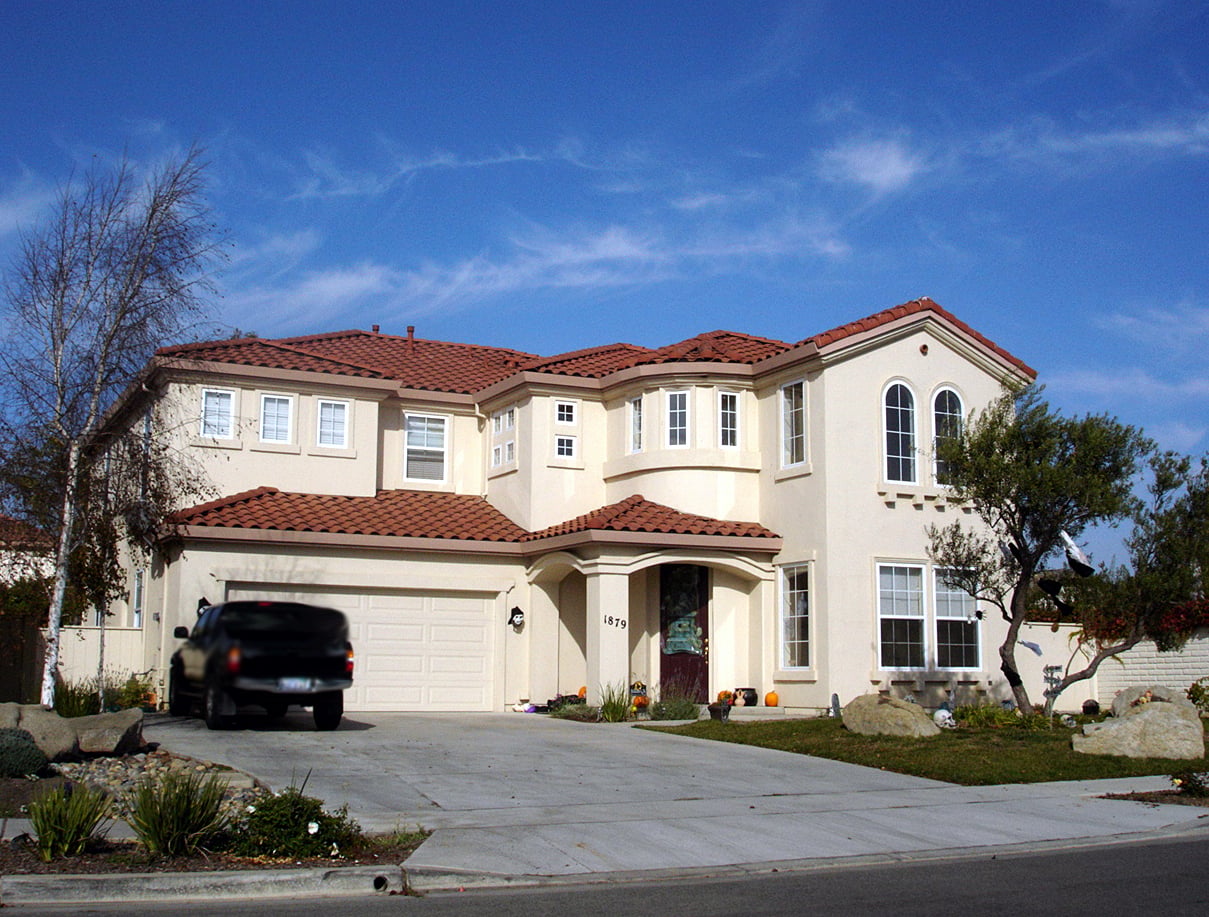 A two-story beige house with a red tile roof, large windows, and a double garage. A black SUV is parked in the driveway, and there are small trees and landscaping in the front yard under a blue sky.