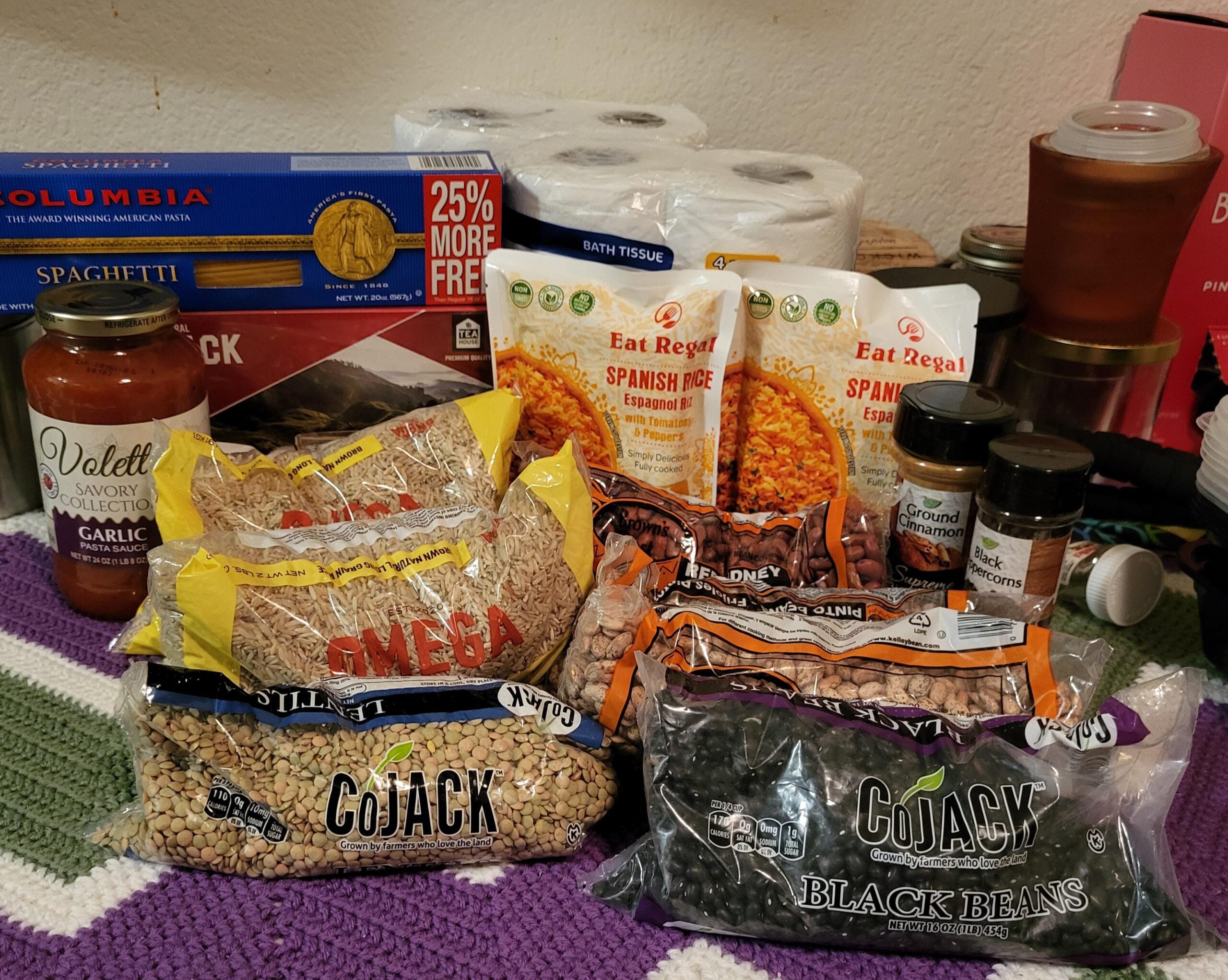 A collection of pantry items on a table, including pasta, spaghetti sauce, lentils, black beans, Spanish rice, various spices, bath tissue rolls, and a jar with a red lid, all displayed on a crochet blanket.