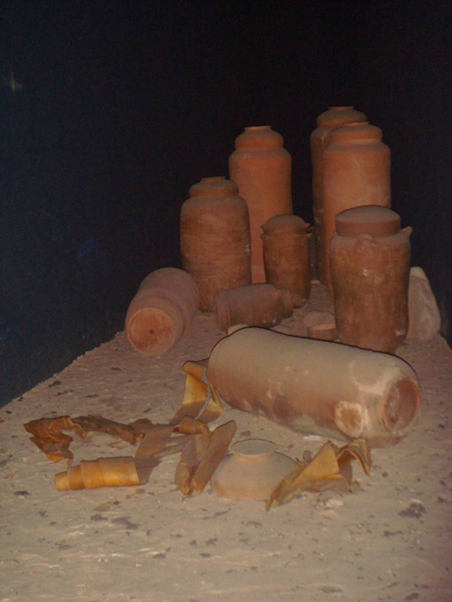 Several ancient clay jars and broken pottery pieces are scattered on a sandy ground, with some rolled-up scrolls lying nearby, all displayed against a dark background.