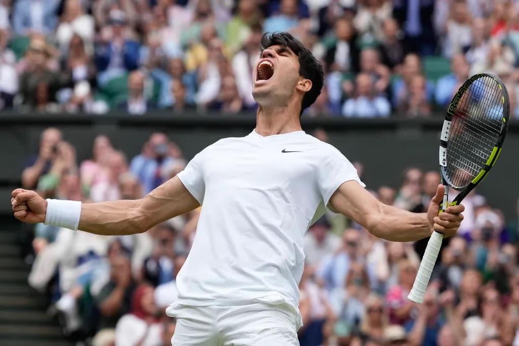 A male tennis player in a white outfit celebrates passionately on the court, clenching his fists and yelling, with a crowd of spectators blurred in the background.