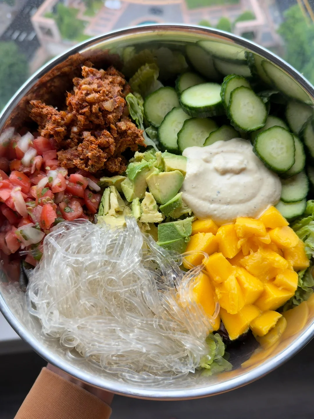 A bowl filled with sections of chopped cucumbers, diced tomatoes, avocado chunks, mango pieces, glass noodles, crumbled plant-based protein, and a dollop of creamy dressing, placed near a window with an urban view outside.