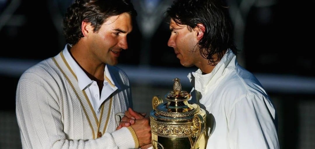 Two male tennis players stand close, shaking hands and holding a large gold trophy, both wearing white outfits. One player looks emotional, capturing a moment of mutual respect after a match.