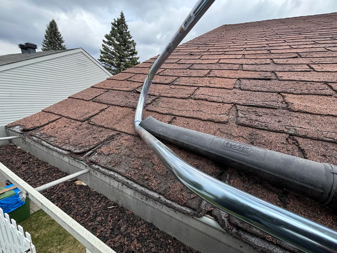 A close-up of a house roof with brown shingles, some debris in the gutter, and a metal pole and hose, possibly for cleaning the gutters. The sky is overcast, and nearby trees and buildings are visible.
