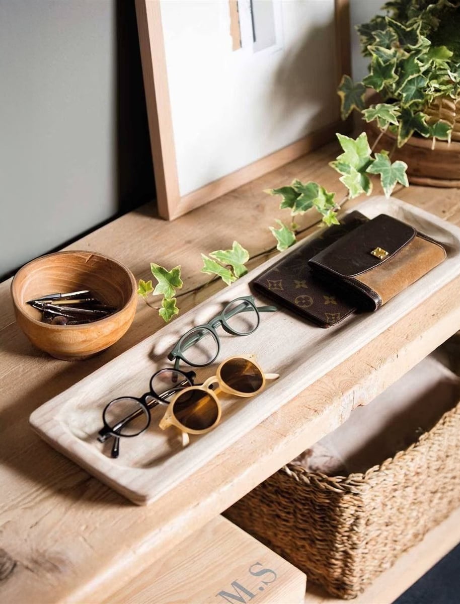 A wooden tray on a table holds three pairs of eyeglasses, a brown wallet, and a pen. Next to it is a small wooden bowl with keys and a plant with green ivy leaves. A wicker basket is underneath the table.