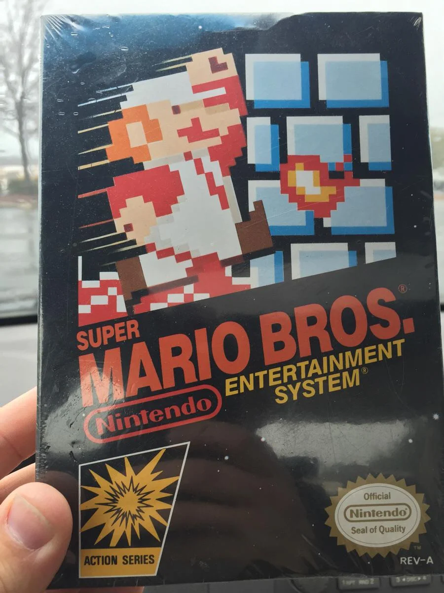 A hand holds a sealed copy of the Super Mario Bros. Nintendo Entertainment System (NES) video game, featuring pixel art of Mario jumping with a brick wall in the background and classic NES branding on the box.