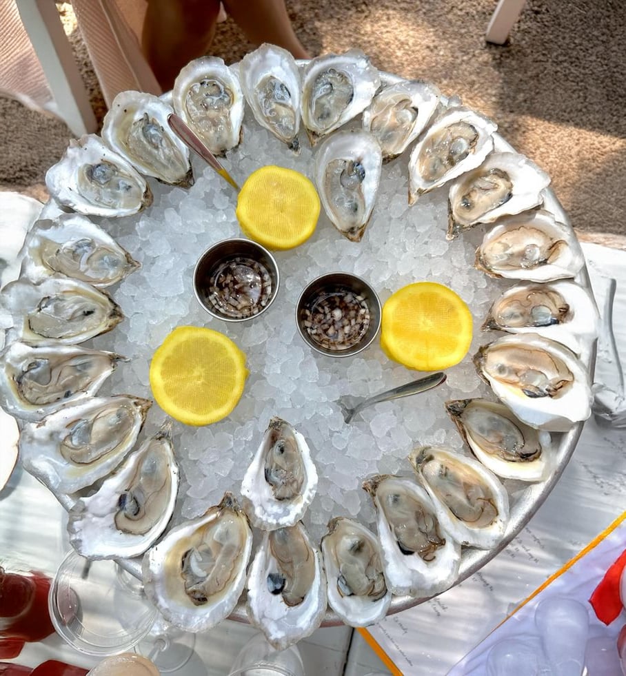 A round platter of oysters on ice, garnished with two lemon halves and accompanied by two small containers of sauce, served with spoons. The platter sits on a white table.