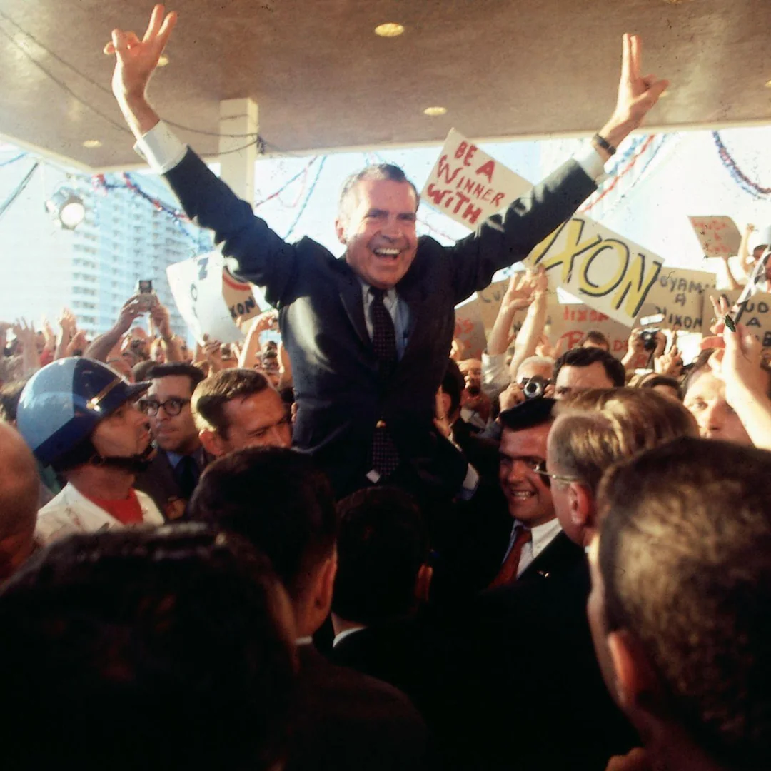 Richard Nixon, smiling and raising both arms in a victory gesture, is lifted by a cheering crowd at a campaign event. Supporters hold signs and cameras, and police are present among the enthusiastic throng.