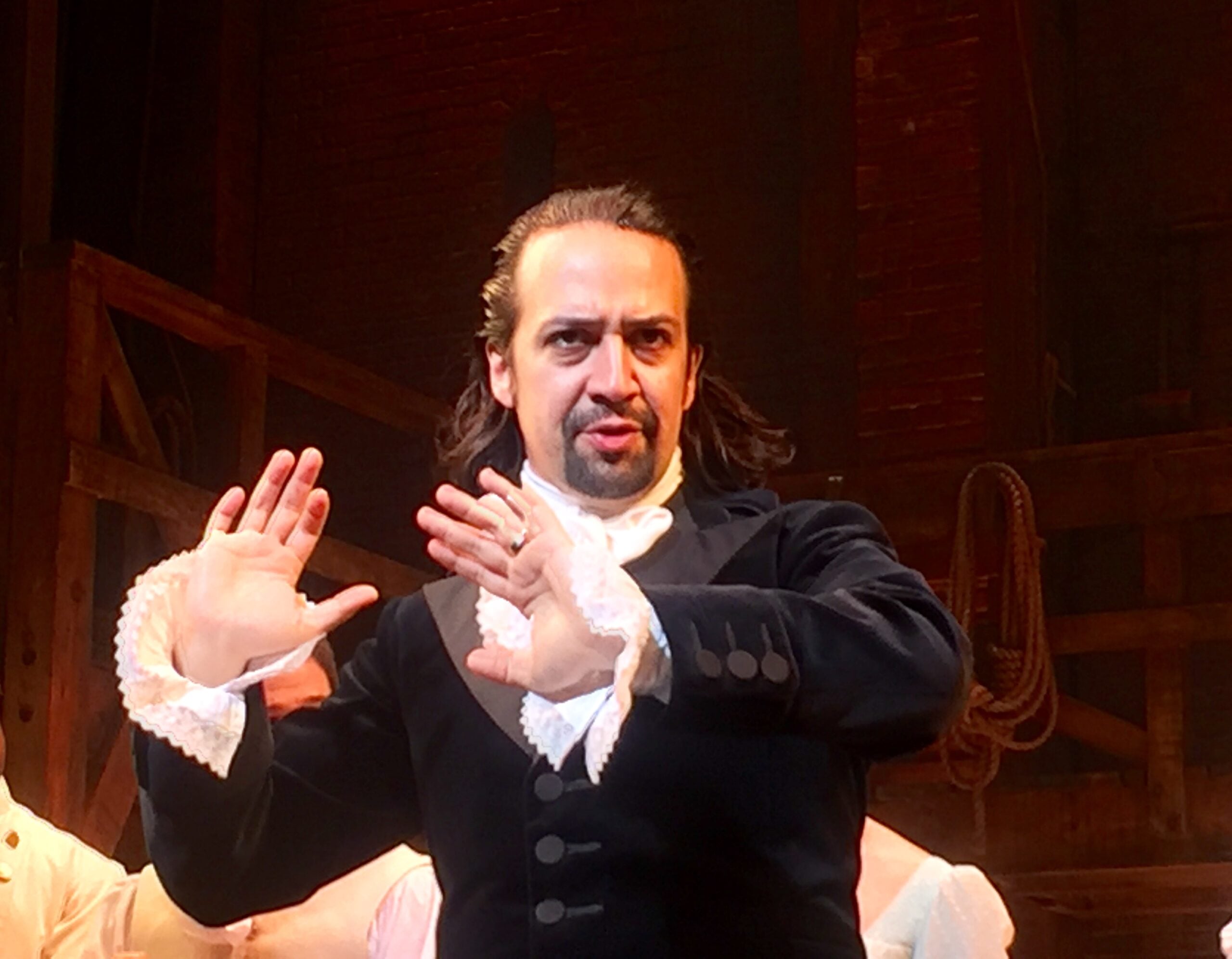 A man in historical clothing with lace cuffs gestures with both hands raised and a serious expression, standing on a dimly lit stage with wooden beams and ropes in the background.