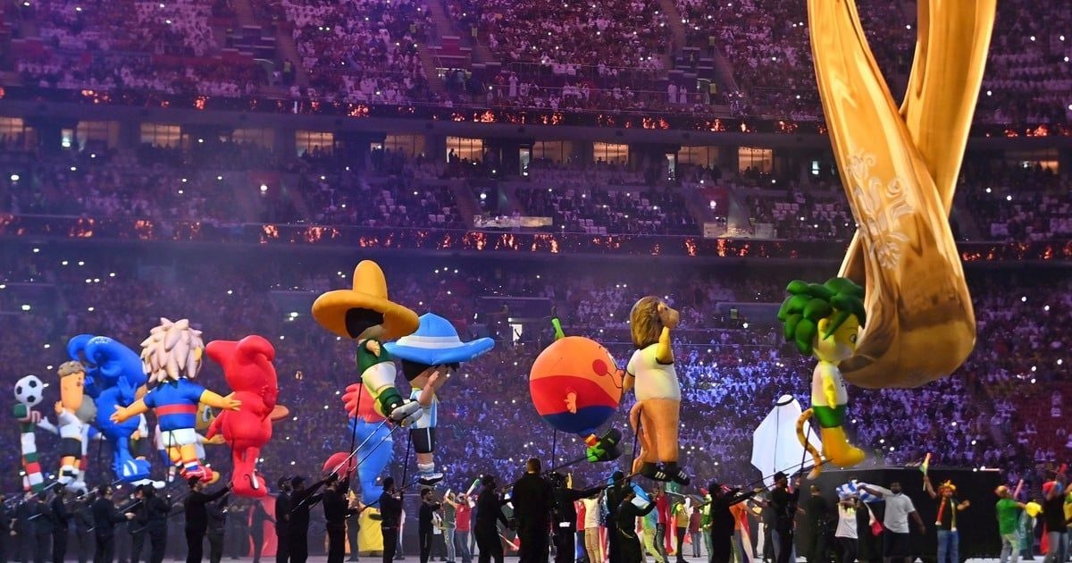 Giant inflatable mascots are displayed in a brightly lit stadium filled with spectators during a festive event, with performers in black outfits guiding the inflatables on the field.