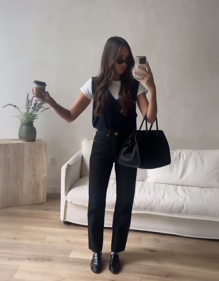 A woman stands in a minimal living room, wearing sunglasses, a white t-shirt, black vest, black pants, and black shoes. She holds a coffee cup in one hand and a black handbag in the other, taking a mirror selfie.