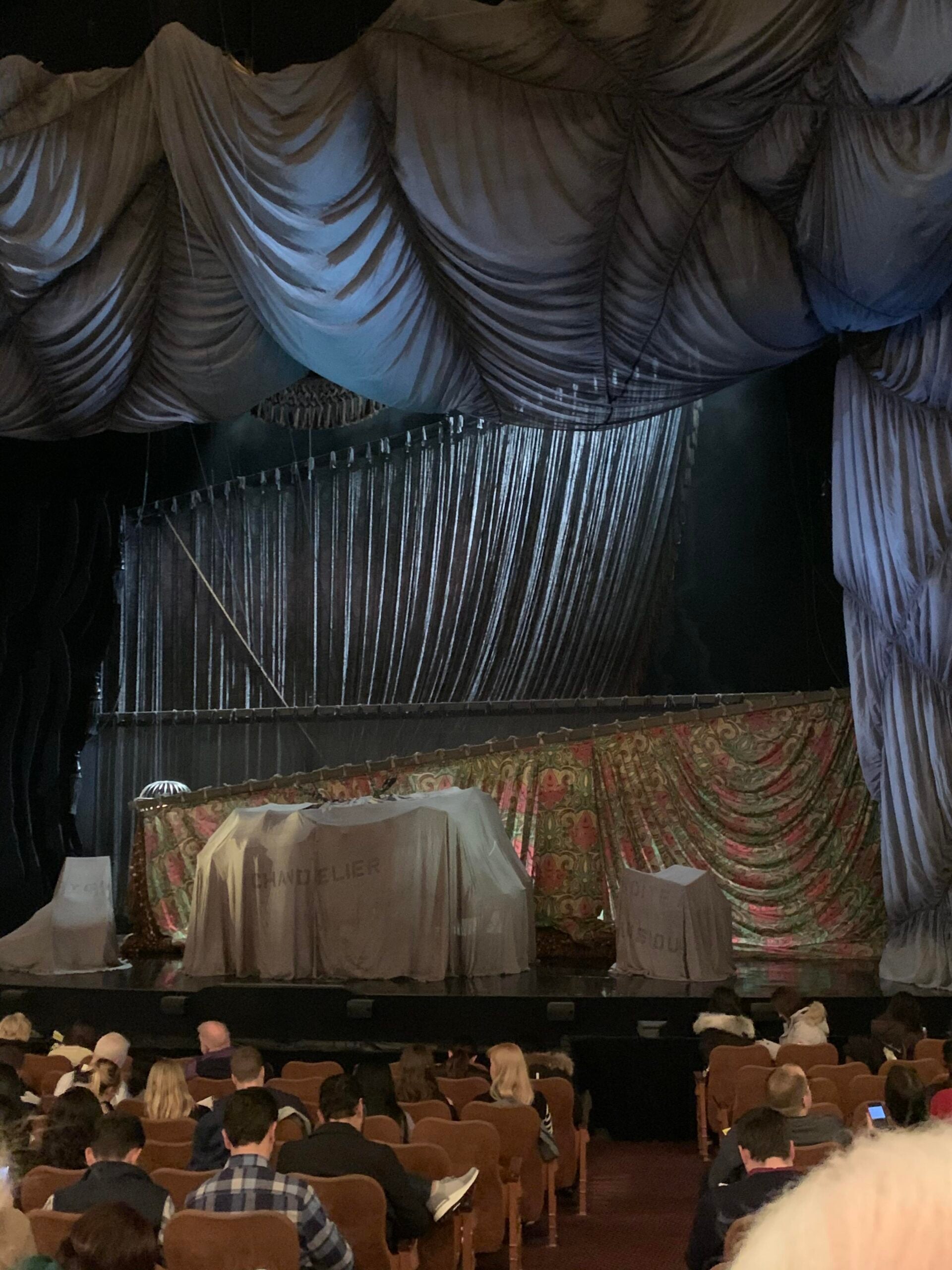 An ornate theater stage with draped curtains and various objects covered in cloth, including ones labeled "chandelier" and "poulie," as an audience waits for a performance to begin.
