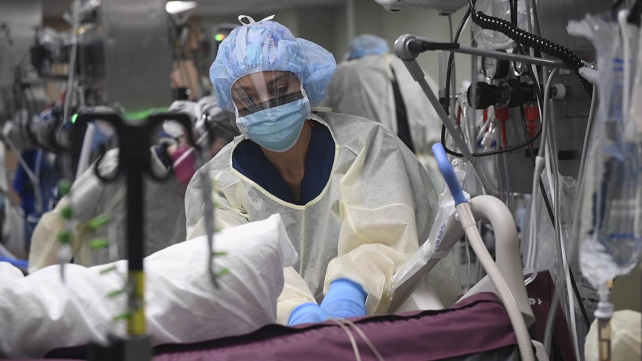 A healthcare worker in protective gear, including a mask, face shield, gown, and gloves, cares for a patient in a hospital room surrounded by medical equipment.